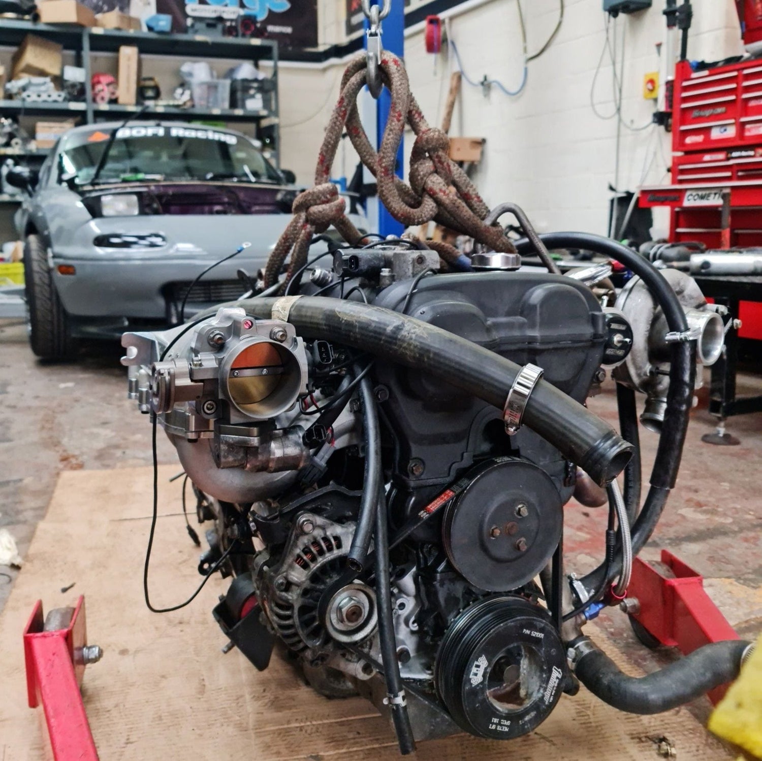 Engine hanging of the engine crane in front of the mk1 Mazda mx-5. Showing resealed engine with all of the ancillaries mounted and ready to be fitted back in the vehicle.