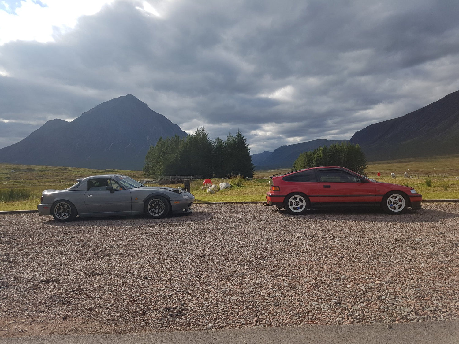 Mazda Mx-5 and Honda Crx parked in front of the mountains in Scotland, Glencoe. Nardo grey mx-5 with wide wheels, low pro headlights, hardtop, front splitter. Crx is in bright red colour, white rota wheels, tinted windows. Both cars are lowered. Stunning mountains in the background.