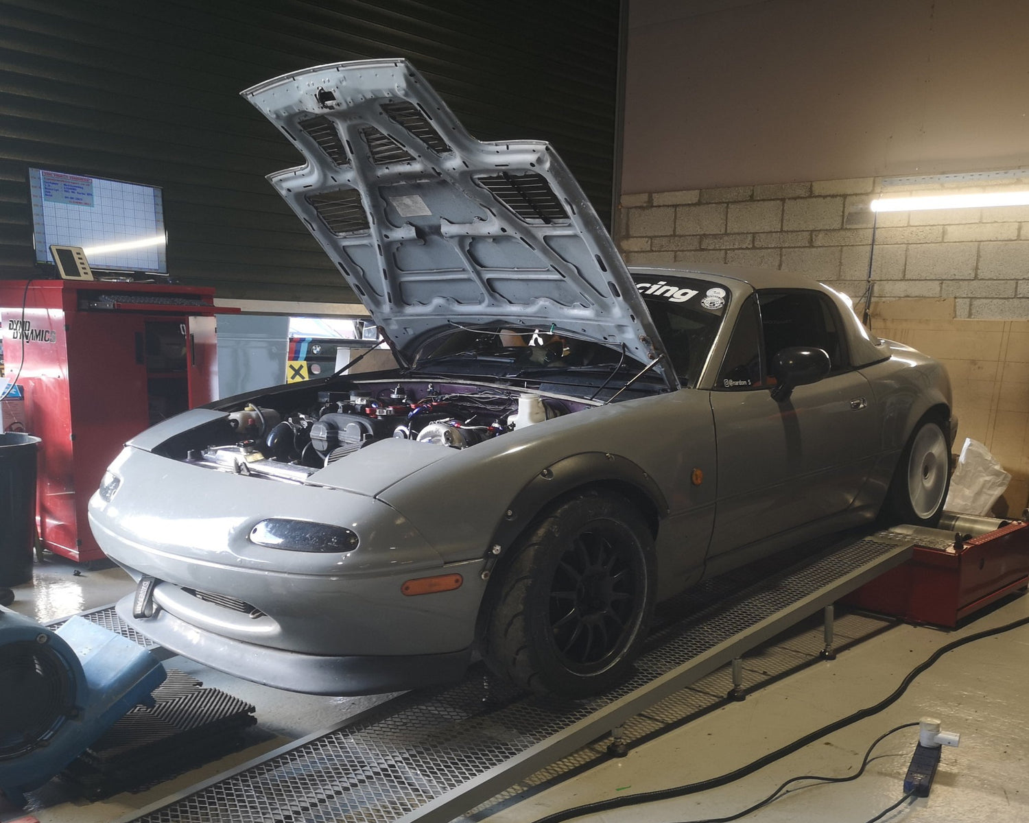 Grey Mazda mx-5 on the rolling road - DYNO, performing a power run. Rear wheel are pining, bonnet is lifted up, vehicle is strapped in place.