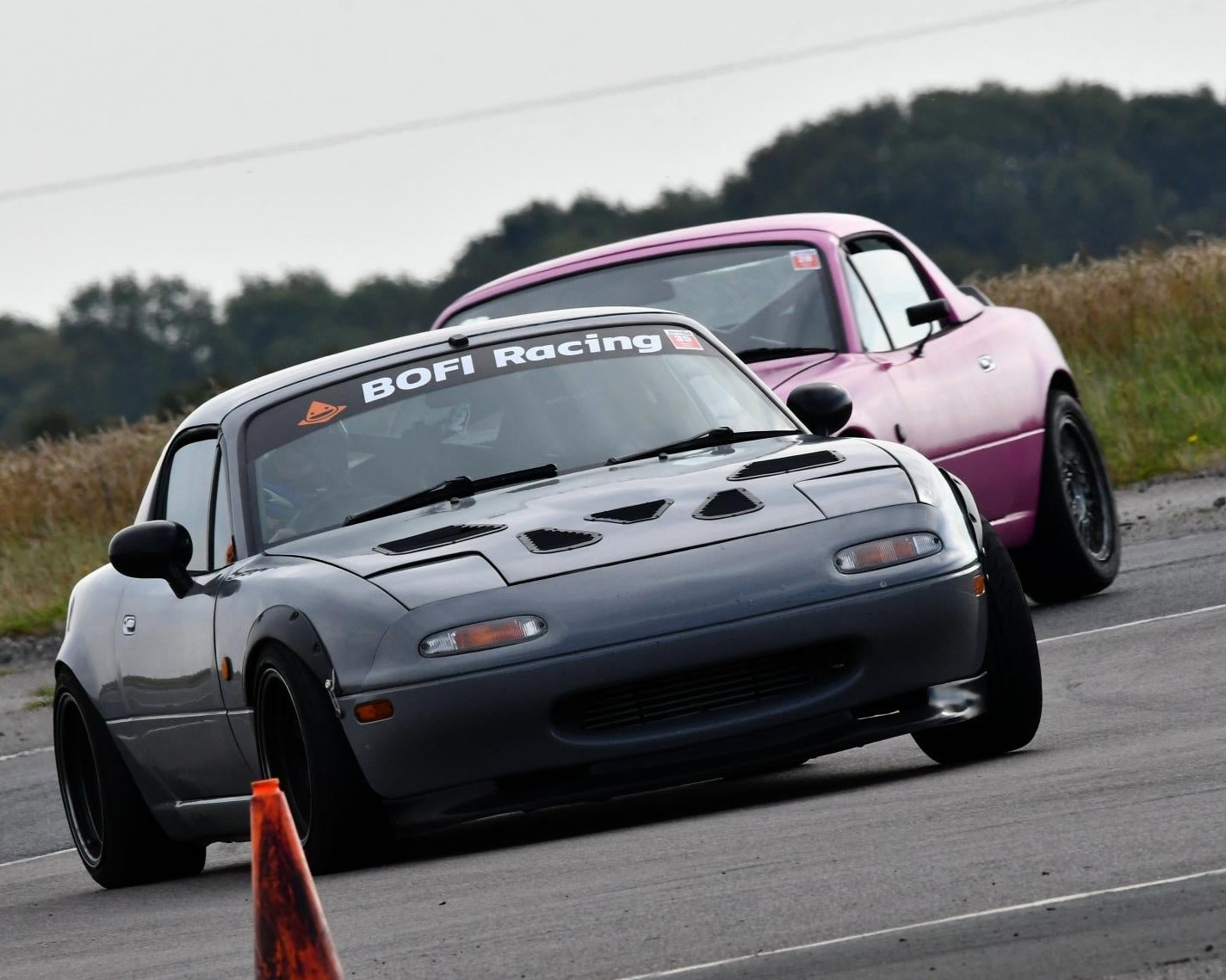 Mazda mx-5 driving at speed on track. Nardo grey, wide arch, lowered, with bonnet vents and abig sunstrip with Bofi Racing logo. Behind it, is another mk1 mx-5 in bright pink colour.