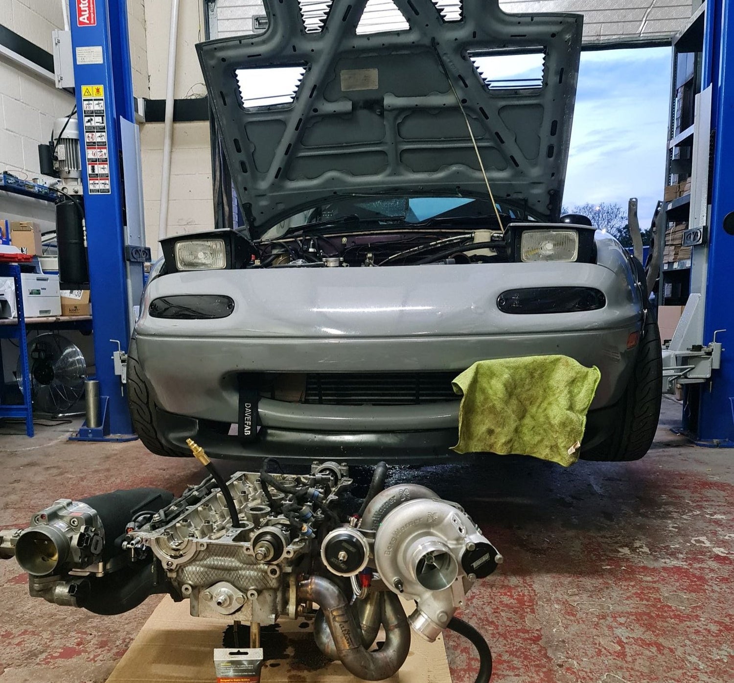 Grey Mazda Mx-5 slightly lifted on the blue ramp. In front of it is engine head stripped into pieces, but with the turbo manifold, turbo and air intake attached.