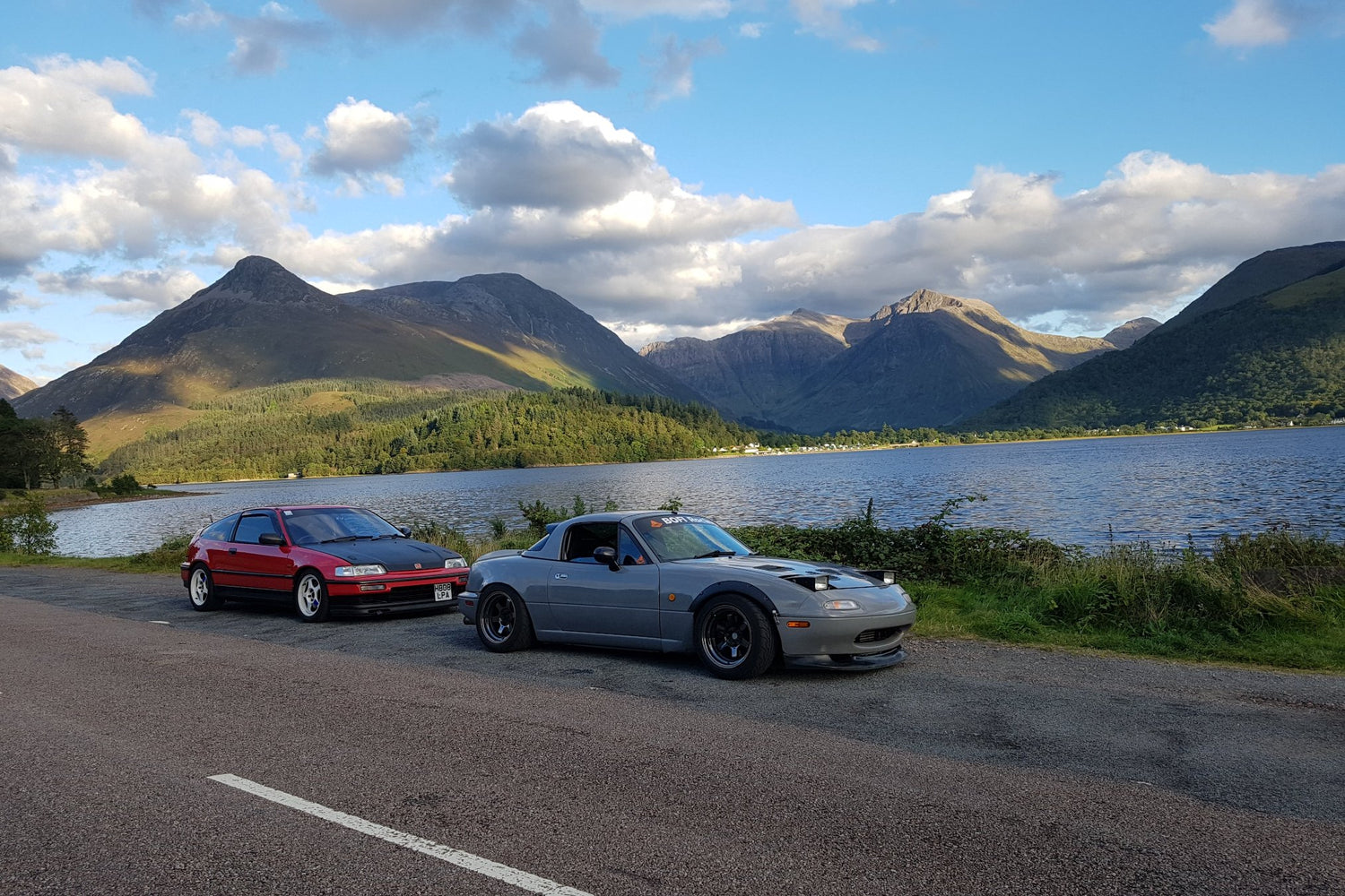 Honda CRX & Mazda mx-5 parked in a layby next to the loch, with a background of the Glencoe mountains in Scottish Highlands. Red CRX with white wheels and black bonnet, Grey mx-5 with wide arches, wide gunmetal grey wheels, low pro headlights, front splitter, hardtop with a spoiler. 