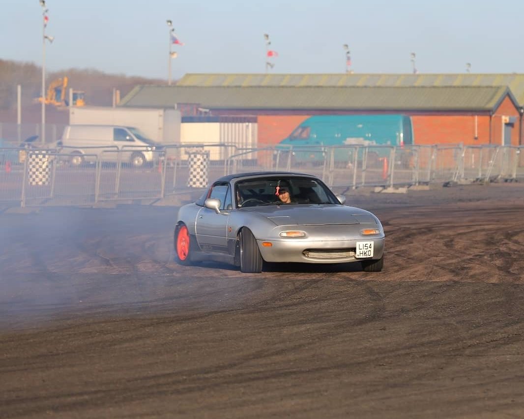 Silver Mazda mx-5 drifting at Santa Pod. Smoke is visible pouring from the tyres, rear wheels are bright pink colour, mx-5 going sideways. In  the background a few buildings, vehicles and a fencing is visible.