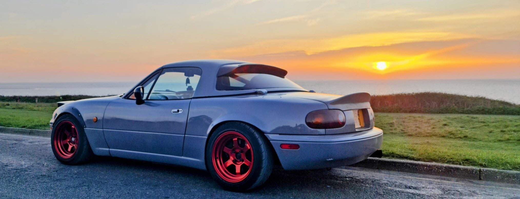 Mazda Mx-5 parked on the cliff top. The sea and the sunset in the background. Mx-5 is in grey colour, with wider apple candy red wheels, two spoilers at the back and wide arches.