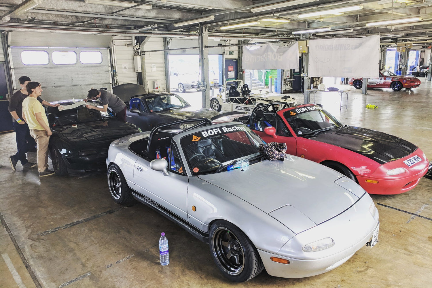 4 Mx-5s inside the pit garage. 3 people fitting a sunstrip on the black mx-5, which is parked at the back next to the blue one. Silver one is in front, and the red on is next to it. There is a kit car in a background.