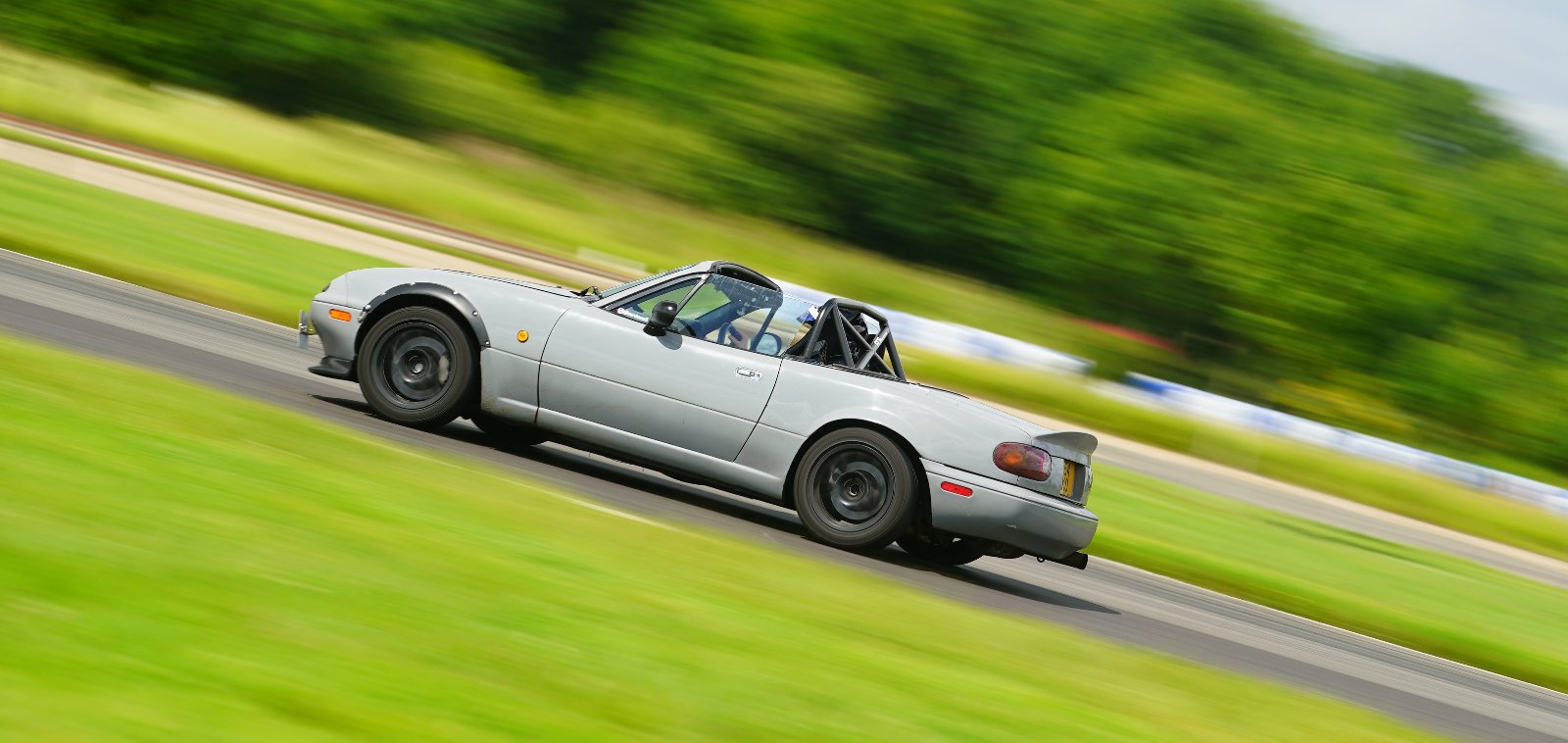 Side shoot of Nardo grey MX-5 on a trackday at Blyton. Very sunny day, blurry bright green background showing trees and grass. Mazda going at a speed. Mx-5 has a ducktail spoiler, wide arches, black wheels, a rollcage and a front splitter.