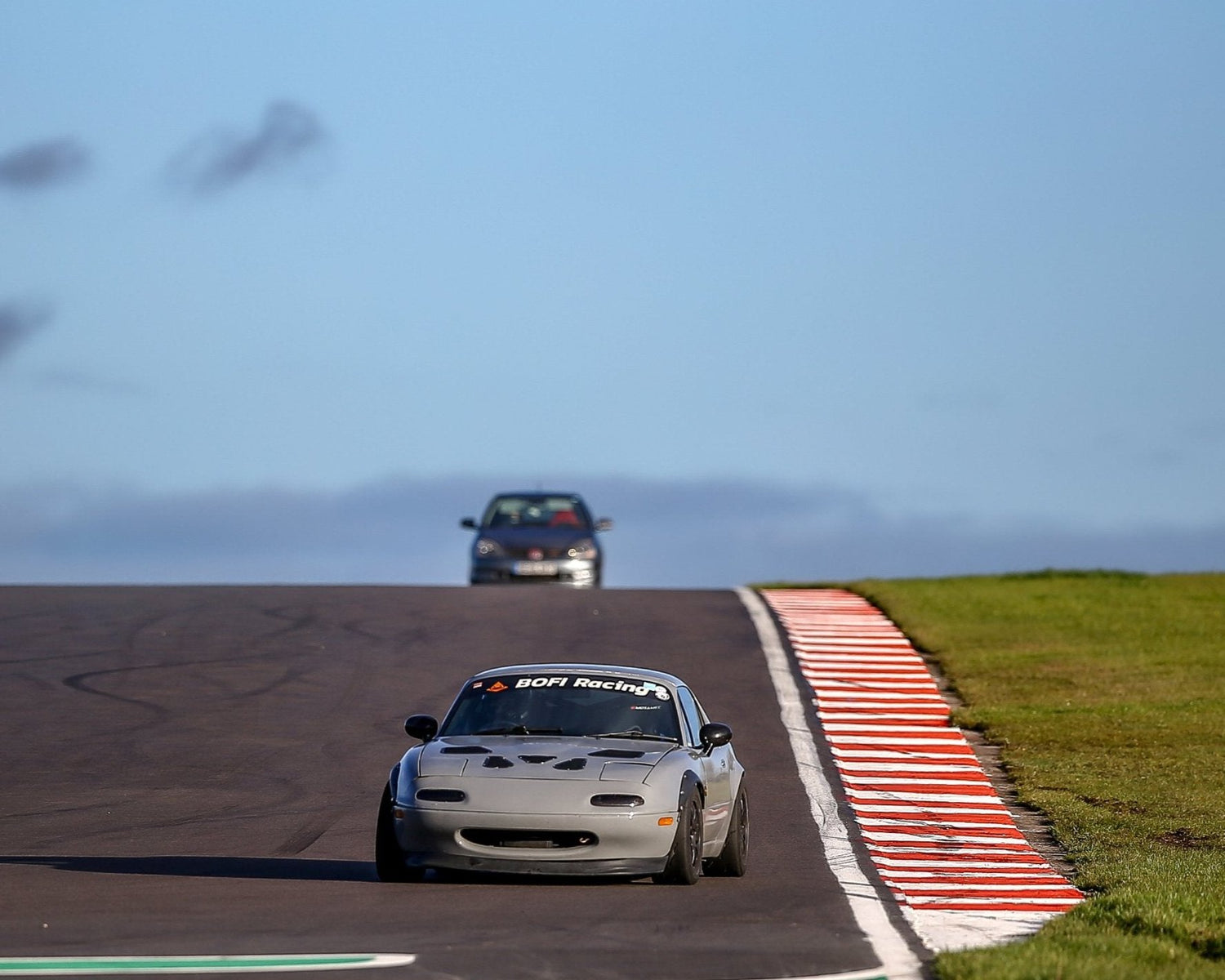 Mazda mx-5 approaching the corner, behind it in a background there is a Honda EP3. Mx-5 is in grey colour, has a sunstrip with Bofi Racing logo, wide arches, black wheels, splitter, bonnet vents.