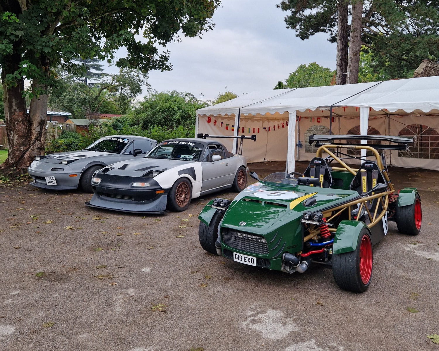 3 vehicles parked in front of marquee. Two mx-5s are heavily modified, one of them having a massive wing and a black splitter. In the front is Exocet in dark green colour, with candy apple red rota wheels and a gold colour cage.